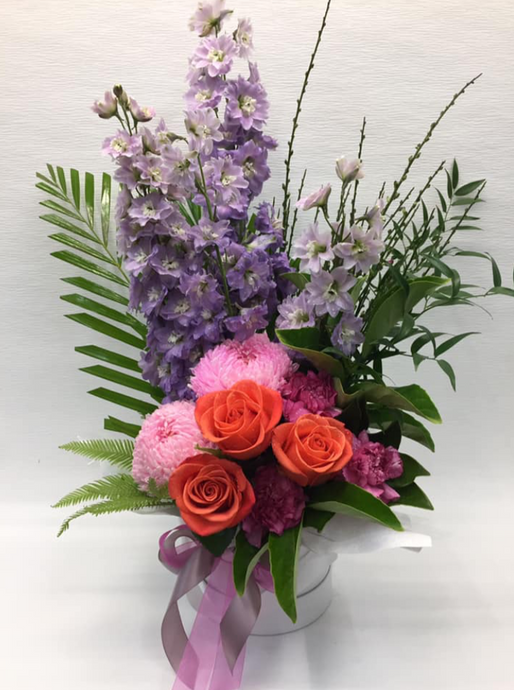 Box arrangement with tall delphinium(when in season) bright disbuds and roses and tropical foliage and agorgeous bow.